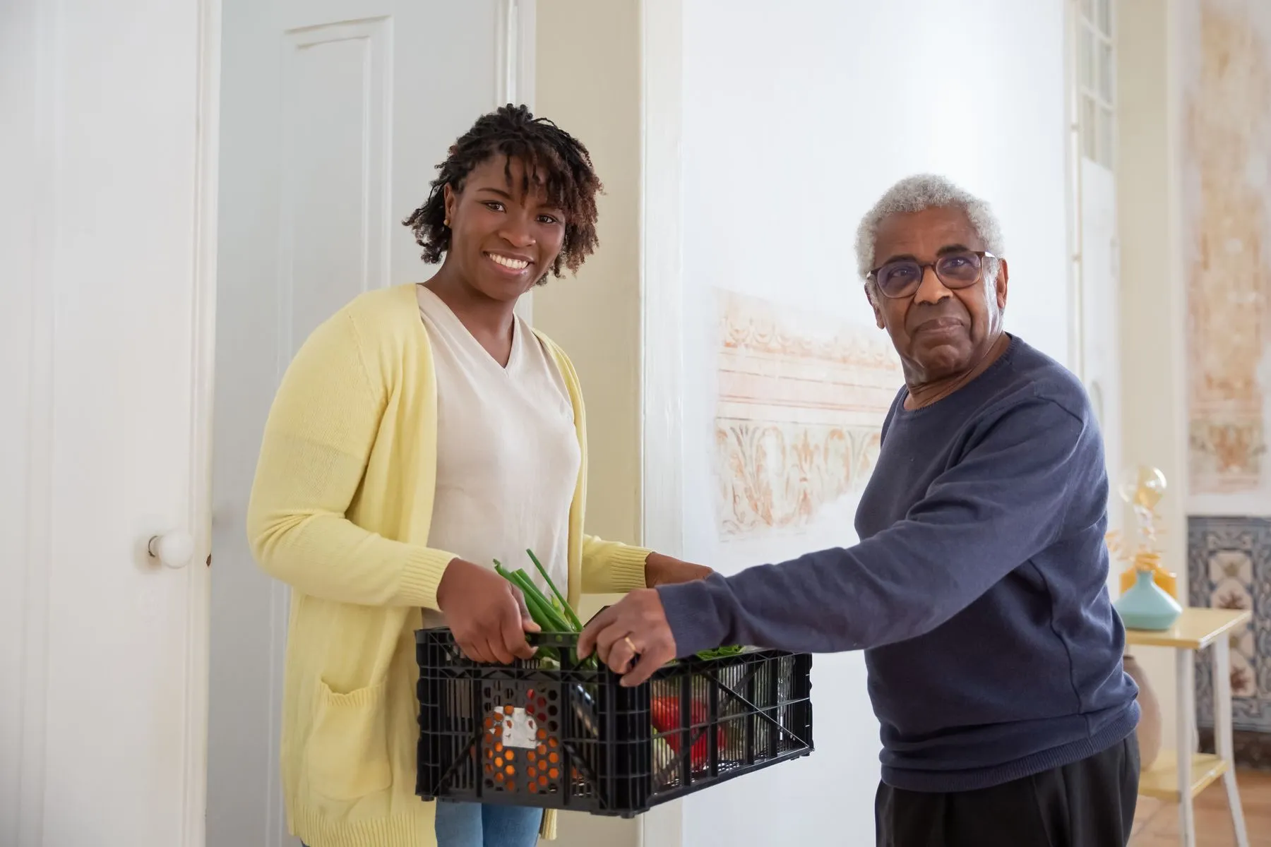 A woman handing a crate of vegetables to an older man at his front door, both smiling