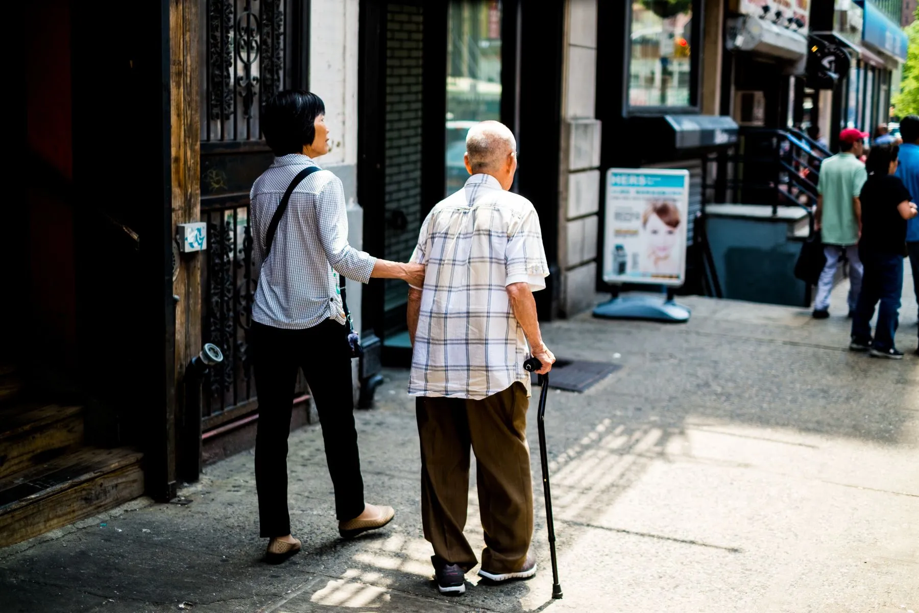 An older man walking along a city street with a walking stick, accompanied by a younger companion